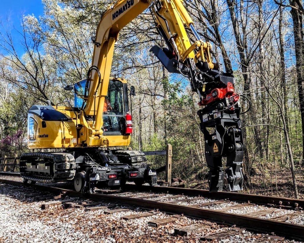 Komatsu 8-ton track excavator on the Dymax Rail Rider 8T hi-rail system fitted with a Rototilt R3 tiltrotator and grapple working a wooded rail right-of-way