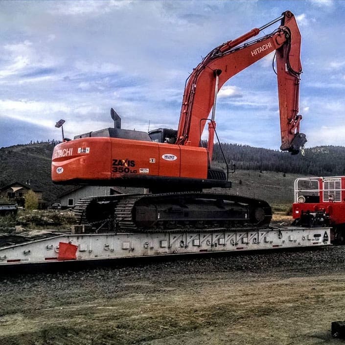 Large excavator being transported on rail by a machinery rail cart and Shuttlewagon railcar mover, with mountains and blue sky in background.