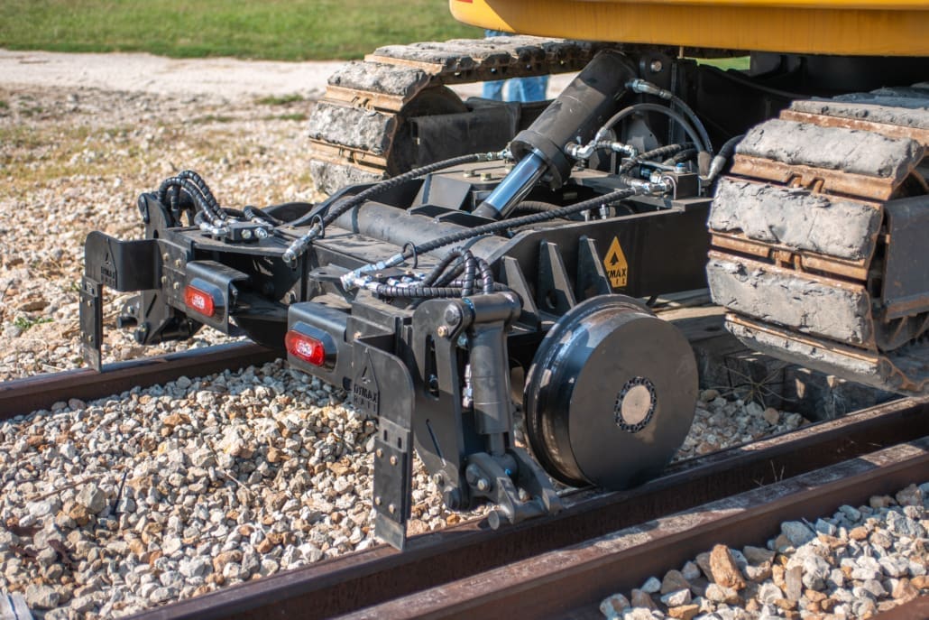 Detail view of the Dymax Rail Rider 8T hi-rail front railgear seated on rail beneath a Cat track excavator, showing rail wheels, hydraulic clamps, and LED lighting modules