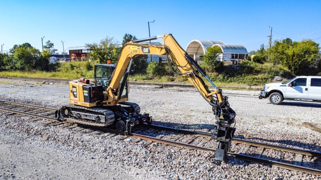 Cat 8-ton track excavator equipped with the Dymax Rail Rider X2-8T hi-rail system working in a railroad maintenance yard