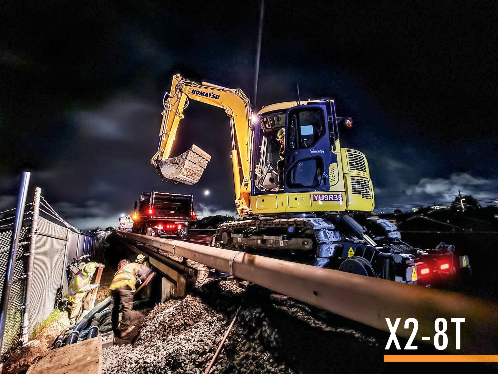 Dymax Rail Rider X2-8T hi-rail system mounted on a Komatsu 8-ton track excavator working a rail line at dusk