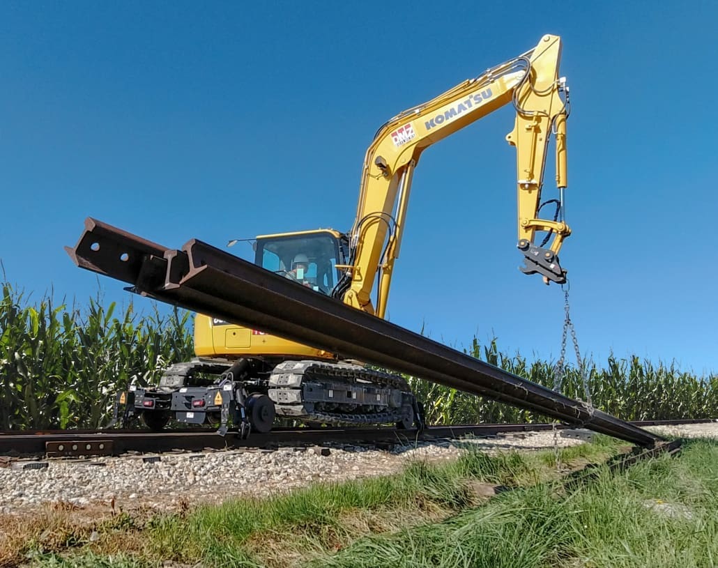 Komatsu 8-ton track excavator on the Dymax Rail Rider X2-8T hi-rail system lifting a length of running rail with chains on a rural rail line bordered by cornfield