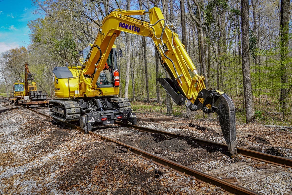Komatsu 8-ton track excavator equipped with the Dymax Rail Rider X2-8T detachable hi-rail system traveling a single-track line through a wooded right-of-way