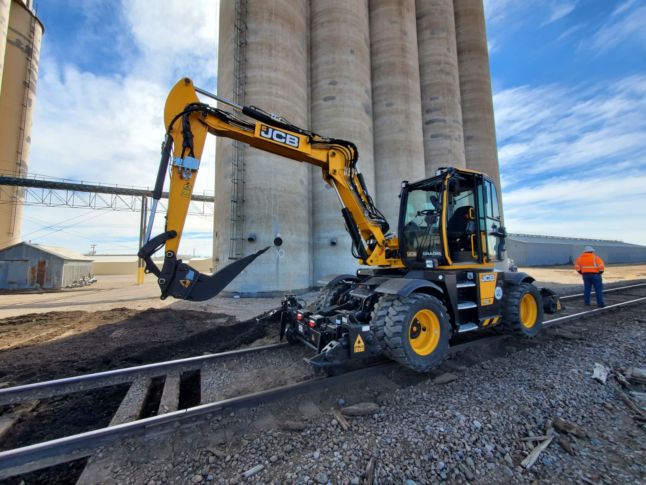 Wheeled excavator with Dymax Rail Rider WX4S hi-rail attachment at railroad crossing Sennebogen 718 wheeled excavator with the Dymax Rail Rider WX4S hi-rail attachment in transport mode at a railroad crossing, showing boom tucked into the railgear with the attachment still on