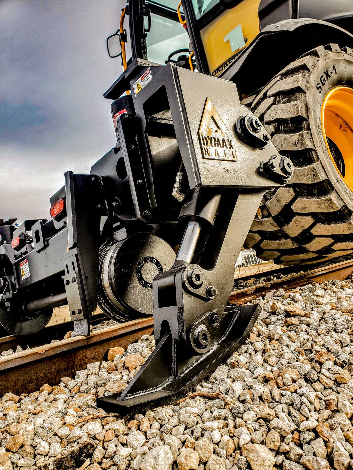 Wheeled excavator with Dymax Rail Rider WX4S hi-rail attachment at railroad crossing Sennebogen 718 wheeled excavator with the Dymax Rail Rider WX4S hi-rail attachment in transport mode at a railroad crossing, showing boom tucked into the railgear with the attachment still on