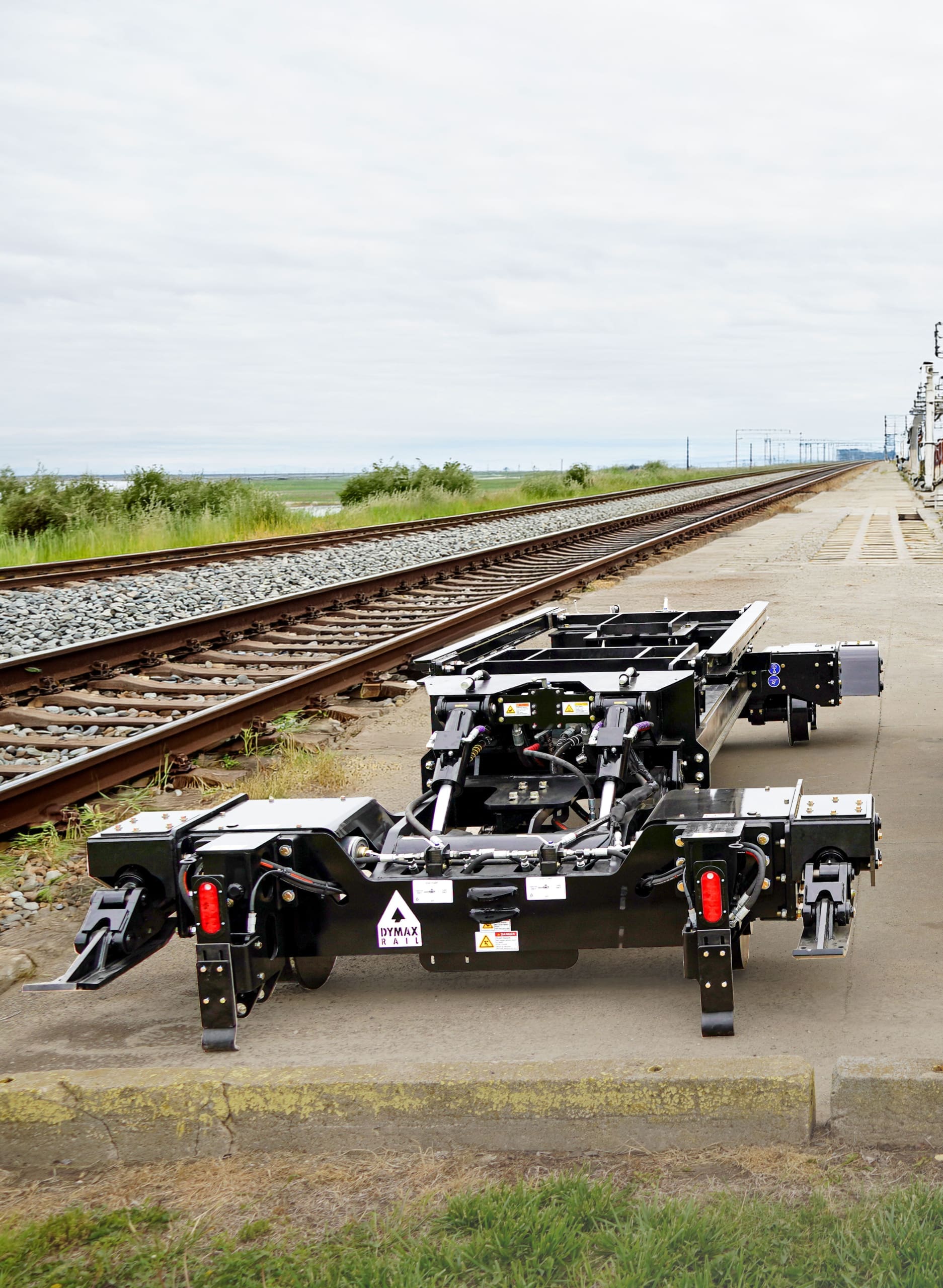 Dymax Rail Rider X2 hi-rail attachment — before installation Dymax Rail Rider X2 detachable hi-rail attachment shown standalone on a concrete pad next to an active rail line, ready for installation on a track excavator